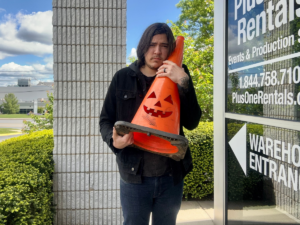 A photo of a man with long brown hair, wearing a black hoodie and blue jeans. He is hugging an orange traffic cone in his arms. The traffic cone has a jack-o-lantern face cut into it. He is standing outside a building, which has the name of the business and contact info on the windows.