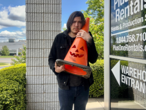 A photo of a man with long brown hair, wearing a black hoodie and blue jeans. He is hugging an orange traffic cone in his arms. The traffic cone has a jack-o-lantern face cut into it. He is standing outside a building, which has the name of the business and contact info on the windows. 