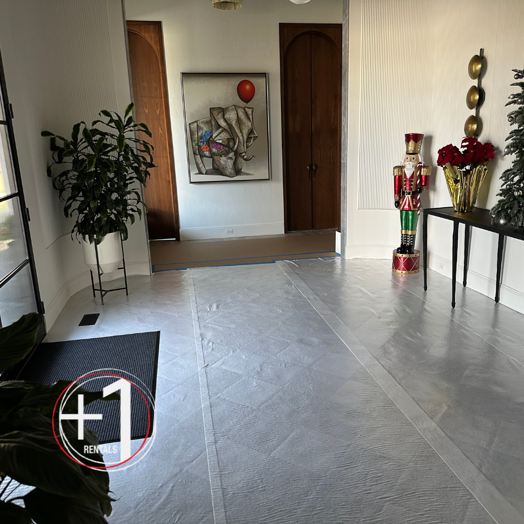 A photo of a room with white flooring protectant covering the floor, a tall plant in the corner, a rubber door mat by the front entrance, and a table with Christmas decorations on the right wall. Film location flooring protection DMV Washington DC Maryland